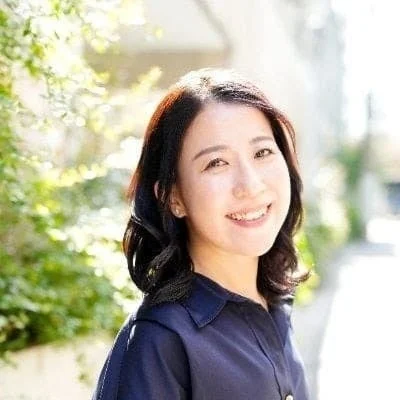 屋外で笑顔を見せるアジア人女性のポートレート。暗い色のシャツを着用し、背景には緑豊かな植物がぼかして写っている。