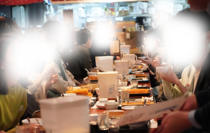 複数の人々がレストランや居酒屋のような場所で長いテーブルを囲んで食事をしている様子