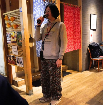 長髪で眼鏡をかけた男性がマイクを持ち、屋内で話しているか歌っている様子