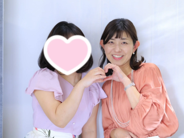 二人の女性が並んでおり、一人は笑顔で両手でハートのポーズを作っている画像