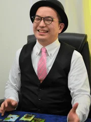 黒いハットと眼鏡をかけ、ピンクのネクタイとベストを着用した男性が笑顔
