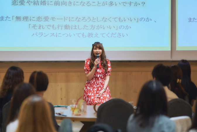 赤いドレスの女性がマイクを持って講演、スクリーンに質問