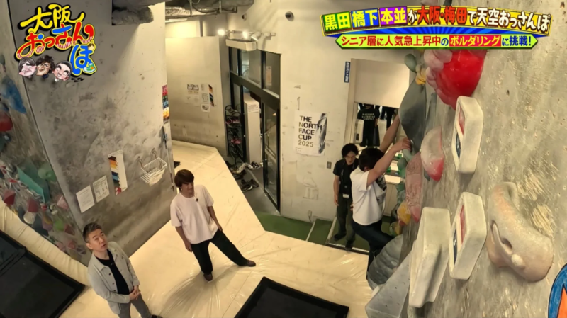 ボルダリングジムで数人の男性がボルダリングに挑戦している様子を捉えた画像です。テレビ番組「大阪おっさんぱ」のロケのようで、出演者がシニア層に人気のボルダリングに挑む様子が映し出されています。