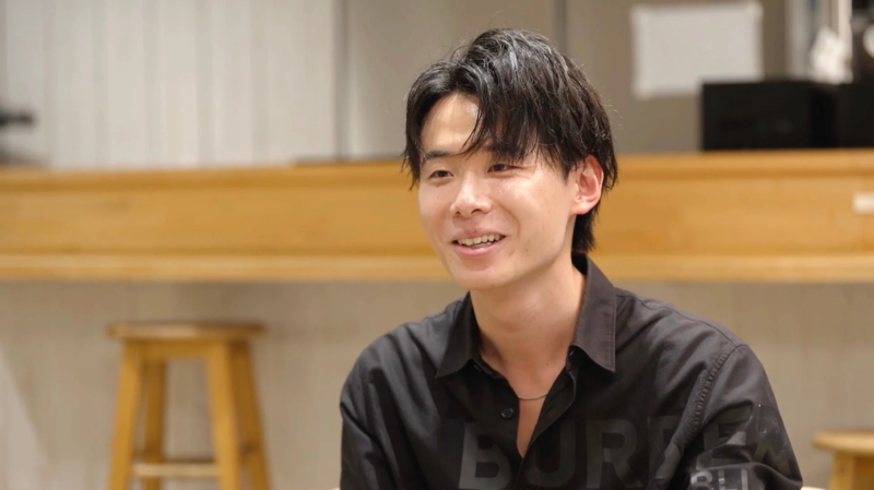 黒いシャツを着た若いアジア人男性が、明るい笑顔で左側を見ています。