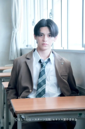 学生服を着た若い男性が教室の机に座り、カメラをまっすぐ見つめている
