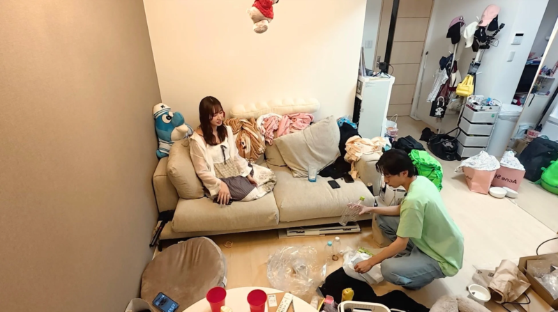 散らかったリビングルームで、女性がソファに座り、男性が床で片付けをしている様子が捉えられています。
