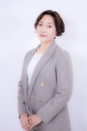 白い背景の前で、グレーのジャケットとパールネックレスを身につけた笑顔の中年女性のポートレート写真です。
