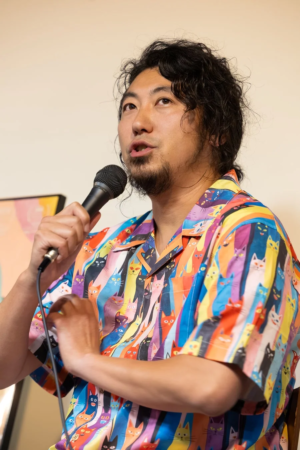 巻き毛と髭の男性が、カラフルな猫柄のシャツを着てマイクを持ち、熱心に話している