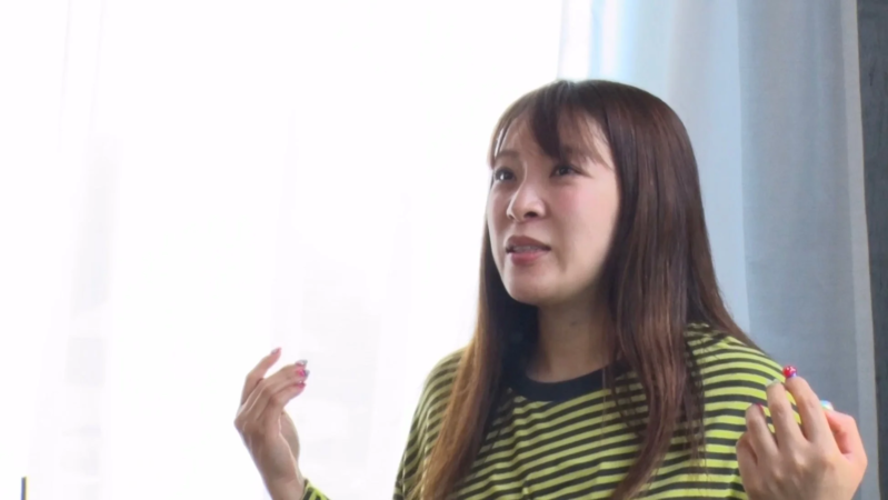 明るい背景の屋内で、ストライプ柄のトップスを着た若い女性が、何かを説明するように手を動かしながら話している様子の画像です。