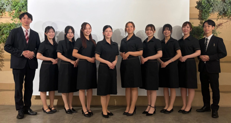 黒い制服とスーツを着用した10名の男女が、白い壁を背景に整列して写る集合写真