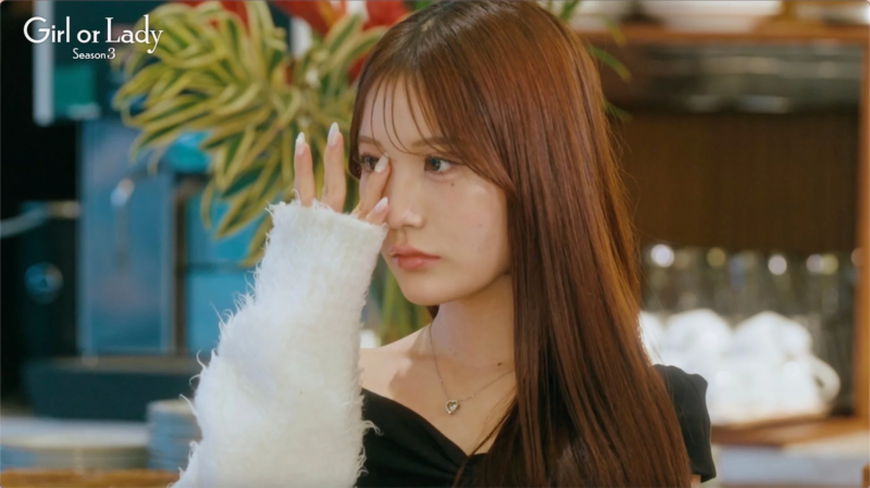 白いふわふわのセーターを着た若い女性が、指で目元を覆いながら何かを考えているような、憂いを帯びた表情を見せている。