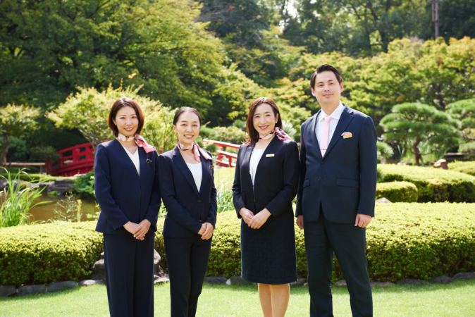 日本庭園を背景に、ビジネススーツと制服を着用した4人のプロフェッショナル(男性1名、女性3名)が笑顔で並んで立っています