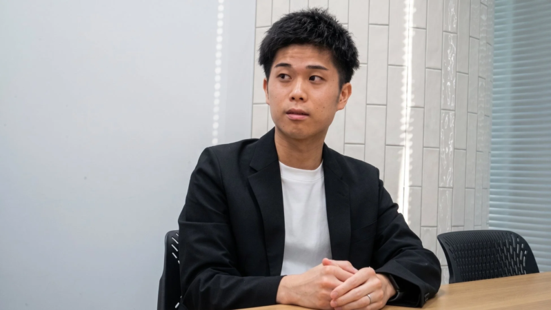 オフィスのような空間で、若い男性がテーブルに座り、真剣な表情で斜め上を見つめている様子。