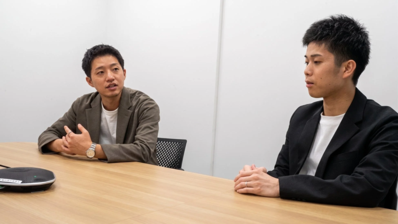 白い壁の部屋で、2人の男性が木製のテーブルを囲んで座り、話し合っている様子。
