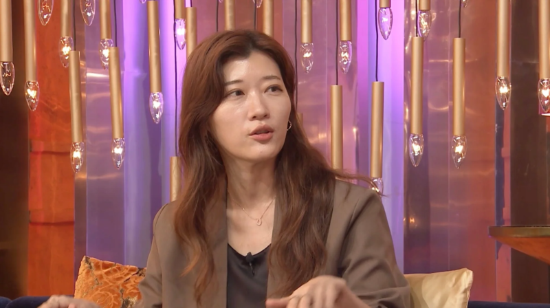 茶色のジャケットを着たアジア人女性が、スタジオのような場所で話している様子