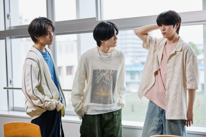 室内で窓際に立つ3人の若者が、それぞれ個性的なカジュアルファッションを着こなしている