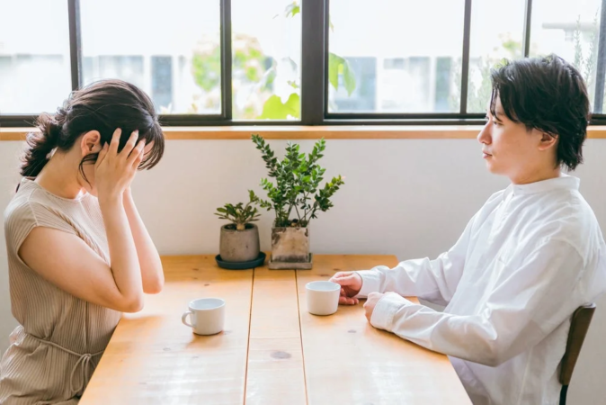 夫婦がテーブルを挟んで座っており、女性は顔を覆って悲しげな様子